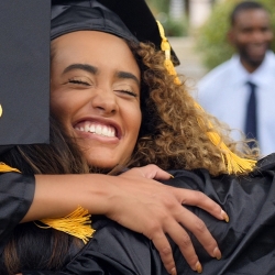 Student in graduation cap and gown hugging another student. Person in button down shirt in back.