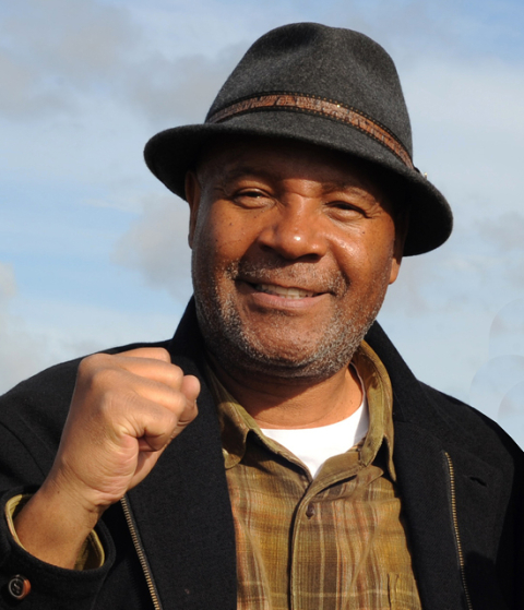 Portrait of artist Emory Douglas wearing a dark hat and jacket, raising a clenched fist.