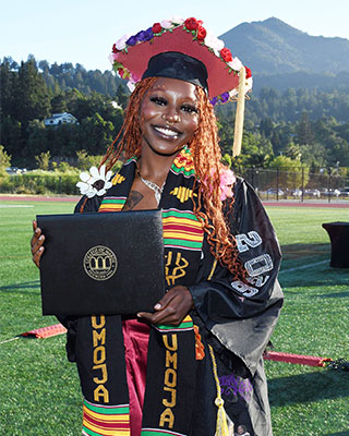 Graduate in cap and gown holding a diploma cover on an outdoor field, wearing a colorful Umoja stole and floral-decorated cap.